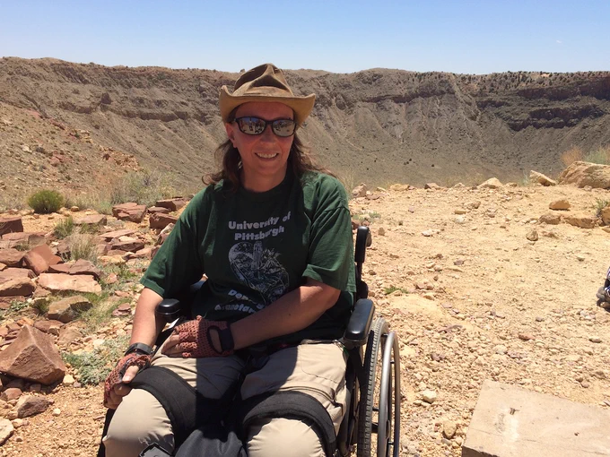 Geologist (with floppy hat, sunglasses, and camera) in wheelchair in front of big hole in the ground (Meteor Crater, AZ)