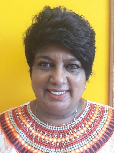 Image shows a women who appears between 40 and 50 years old, with brown skin and dark brown hair smiling directly into the camera. The background is a vibrant yellow.