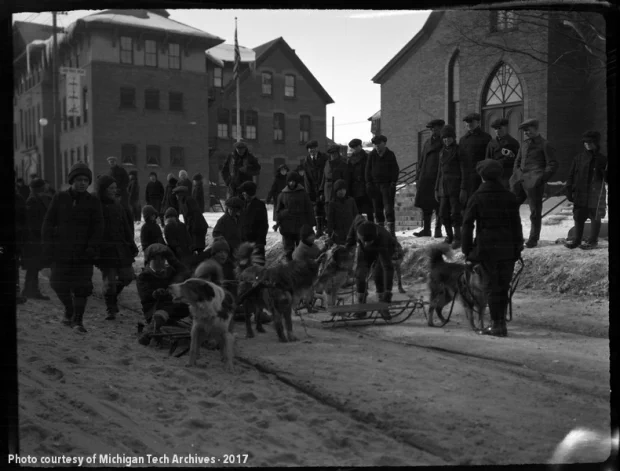 Group of boys and dogs attached to sleds in the middle of a Hancock street