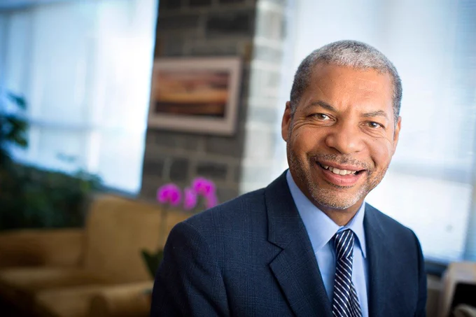 Lance Collins wearing a dark blue suit and striped tie standing in front two windows and purple flowers which are out of focus in the background.
