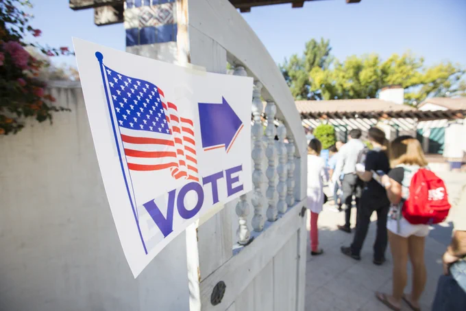A group of students stand in line in front of a red, white and blue vote sign.