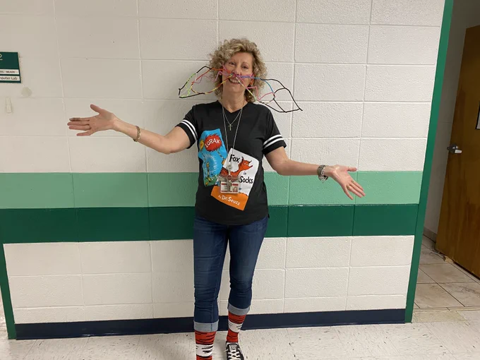 Media Specialist Kathy Sorrell celebrates Read Across America Week. She is wearing a Lorax mustache and a t shirt with Dr. Seuss book covers on it.