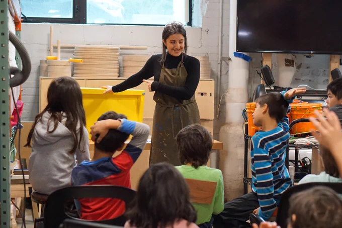 Holding up and pointing to a yellow ramp, Jessica smiles as many of the 8 students hold up their hands to answer a question.