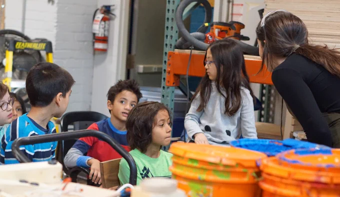 Jessica, our Engagement Coordinator, listens as one of 6 students looks to the side and asks a question in the Makerspace. There are orange buckets of green, red and blue paint in the foreground.