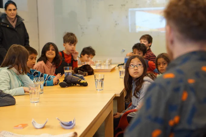 Ten students sit around a wooden table with water glasses. The students are sporting various expressions while one, in a blue striped long-sleeve, is mid-question to Luke, who’s in soft focus in the foreground. Their teacher looks on in the background.