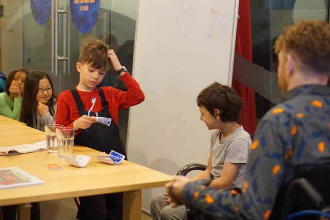 A student wearing a red long sleeve shirt and black overall snow pants, scratches his head while holding a toothbrush and tube of toothpaste. Sitting in front of him is a smiling student in a grey t-shirt, and behind him is a pondering student in a green shirt and a smirking student with a grey long sleeve.