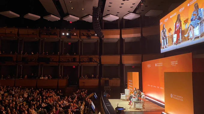 A wide view of the three women on stage with an audience watching.
