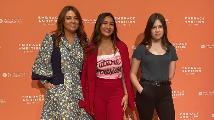 Sam, Deja and Jordan stand with smiles in front of an orange background with the words “Embrace Ambition Summit” many times in white text.