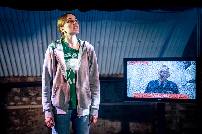 Production photograph from Notch by Steve Gregson. It shows performer Danaja Wass standing on stage next to a tv screen. She stands with her arms by her side looking up and to the left. She wears blue jeans, a green t-shirt with picture of a shamrock on it and a hoodie. On the tv screen is a very blurry image of a man with a moustache, perhaps military, talking.