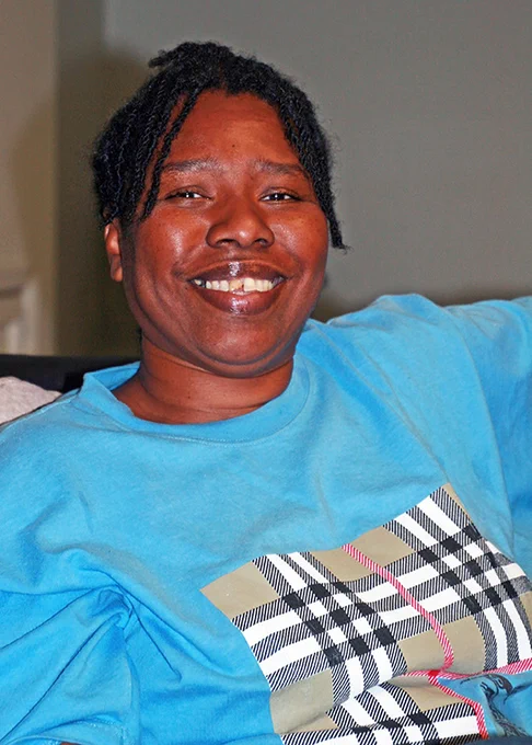 A young Jamaican woman with short corn row braids smiles. She is wearing a blue shirt.