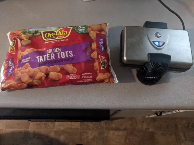 A bag of Ore-Ida brand Golden Tater Tots, next to a plugged-in waffle iron.