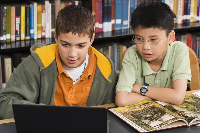 Two boys read from a laptop computer. One boy wears an orange shirt and gray jacket. The other boy wears a green shirt. Behind them is a shelf of books.