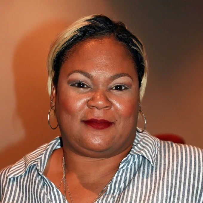 An African American woman with bleached hair smiles. She wears a striped dress.