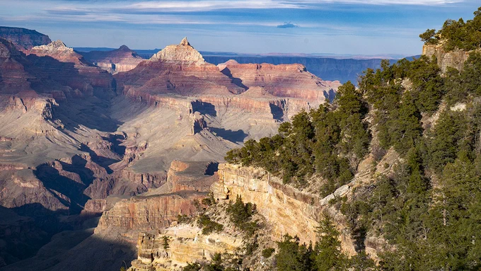 Description: a sloping, tree-covered hillside above a sheer limestone cliff. In the distance, colorful peaks and cliffs rise from the floor of a vast canyon. NPS/M.Quinn