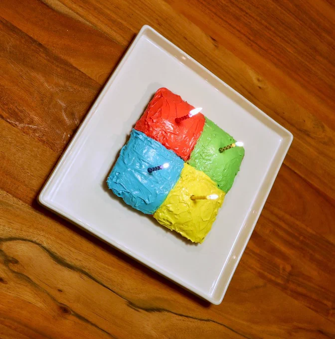 Photography of four differently coloured cakes on a white plate. The plate is resting on a wooden table. Each of the four cakes are coloured to represent the Microsoft logo. A candle is in each cake to celebrate Microsoft's birthday.