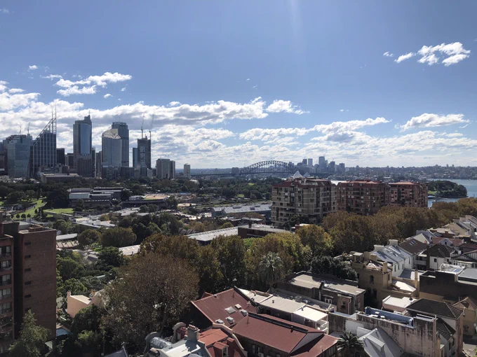 View of Sydney looking northwest from inner east Sydney. From left to right: Sydney CBD (north), Harbour Bridge, North Sydney and partial view of harbour at Woolloomooloo. Apartment complexes and houses in foreground. Weather is sunny with blue sky and some cloud.