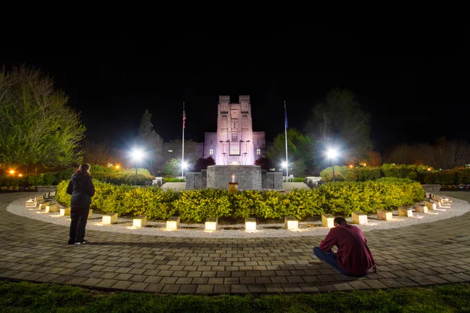 Two Hokies observe the April 16 memorial on the 2020 Day of Remembrance, in the early hours of the morning, with a lit Burruss Hall in the background.