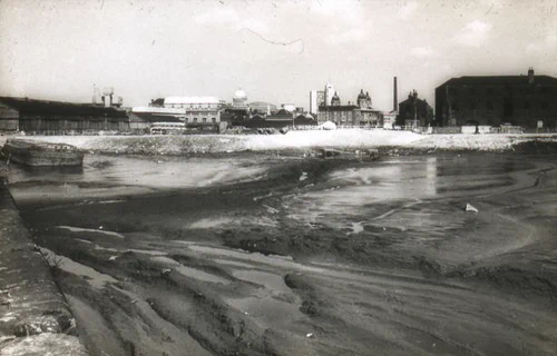 A black and white photograph of a an empty dock that has been filled in with mud. A barge is moored up on the left side of the dock and there are buildings in the background.
