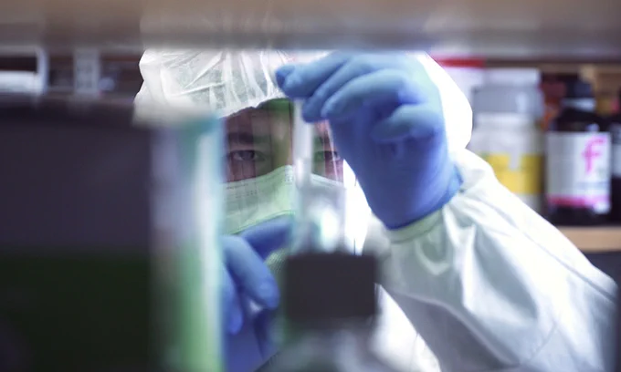 Researcher in mask and face shield holds a test tube.