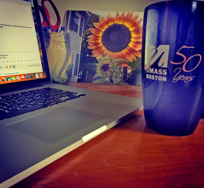 Workspace at home has a laptop and UMass Boston 50 Years mug