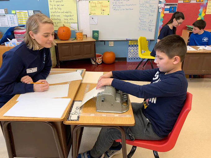 Delta Gamma volunteer assisting a boy with his braille writer