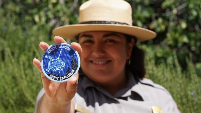 A woman with brown hair wearing a national park service uniform and hat holds up a patch labeled Junior Ranger Night Skies Explorer.