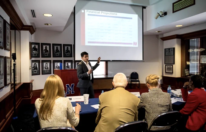 A student in a suit presents before a panel of judges with a Power Point presentation behind him at the University of Mississippi.