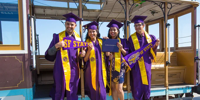 Photo of four SF State graduates at Commencement at Oracle Park standing in a cable car