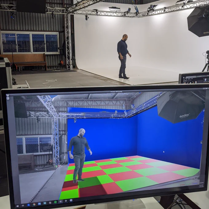 Shot of a computer monitor in the foreground showing a live video feed with graphical overlay. In the background is the live scene with a person in front of a white screen