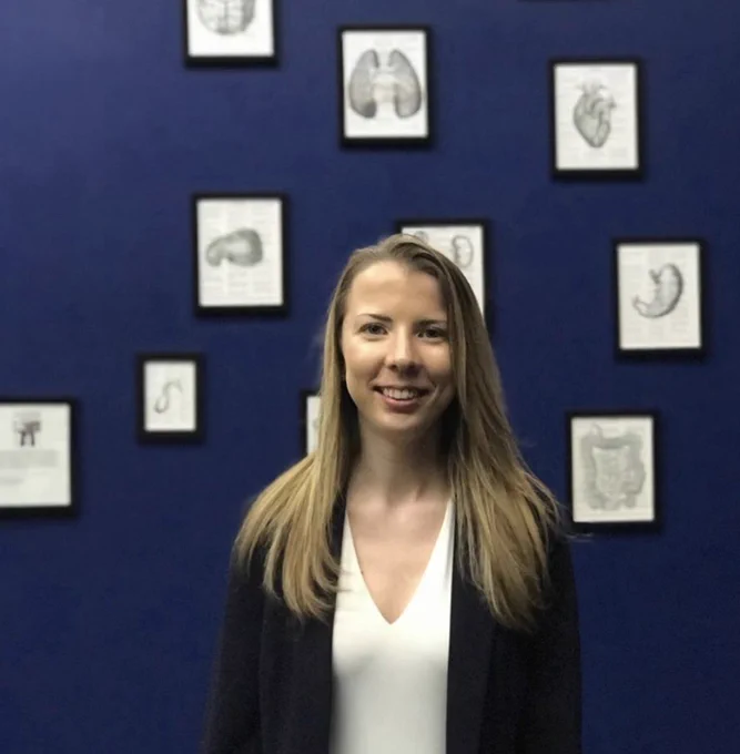 Amanda (white woman with blonde hair) standing in front of a dark blue wall with framed photos of organs she drew hanging from it.