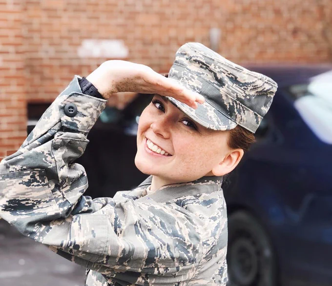 woman standing outside wearing army fatigues, smiling, using her left arm to form the Greek letter Gamma in front of her face