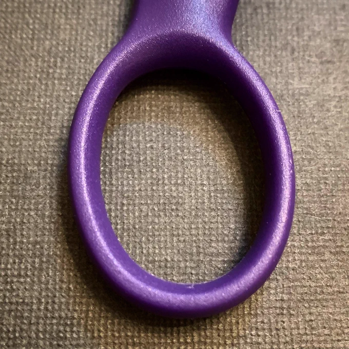 The circle part of purple handled scissors sitting on a textured dark grey background