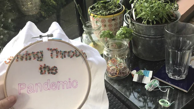 picture of an embroidery hoop with a work in progress spelling out 'Sewing Through the Pandemic' in colourful dots on the left side, and a table with various herbs in pots and embroidery threads on it on the right