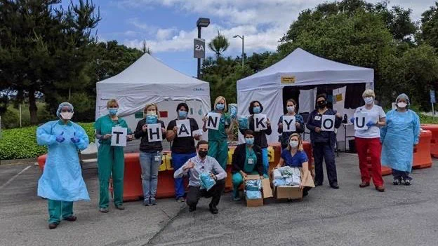 Group of medical professionals stands outside COVID tents with individual letters per person spelling THANK YOU with boxes of PPE