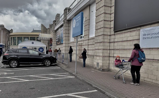 This is a long queue outside Tesco.  Closest to us is a lady with a baby in a park.  It is an overcast day and looks like it might rain..  But hey, its Scotland.