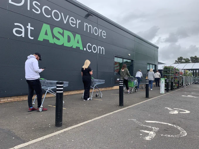 A queue outside Asda. People have trolleys and most are looking at their phones.  The wording on the wall says Discover more at Asda .com.