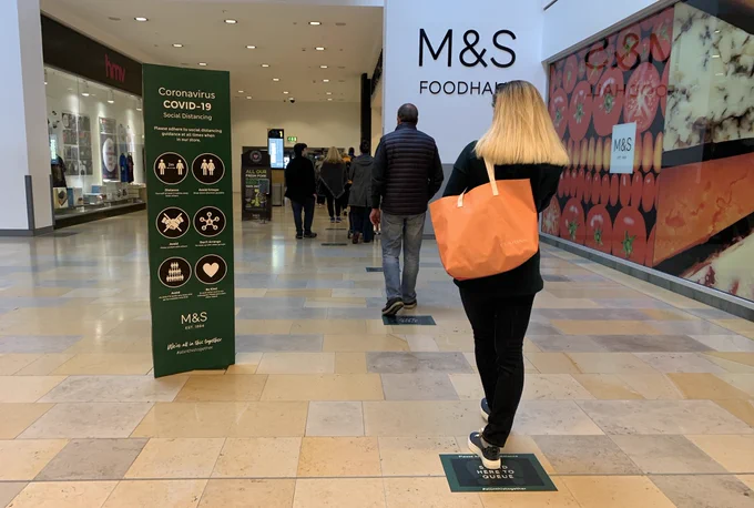 A queue outside Marks and Spencer.  Every one is standing on signs on the floor that ensures people social distance.  There is a banner on the left explaining COVID19 rules.