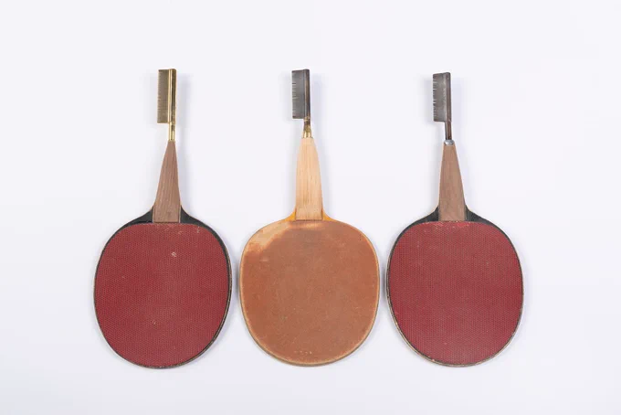 Three tennis paddles arranged in a row and upside down on a white background. The paddles are red and rust colored with wooden handles. Extending from the end of each handle is a black and gold hot comb facing to the left.