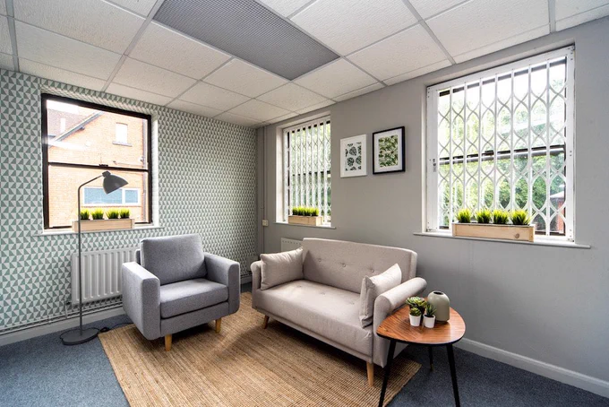 A photo of one of the counselling rooms at Palmer House. There are three large windows. One wall is grey and the other has a green geometric wallpaper. There is blue carpet and a large wood effect rug. There is a grey armchair against one wall and a beige couch against the other wall. To the side of the couch is a small teak side table with 3 small succulents and a vase on it. The room is bright, modern and spacious.
