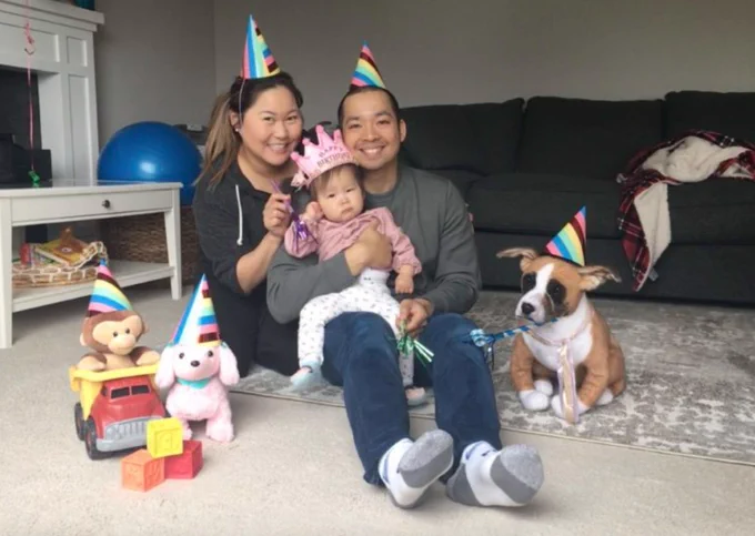 Student Jessica Pak Mortega holds her baby daughter while sitting on living room floor next to her partner, Edwin. They wear birthday hats.