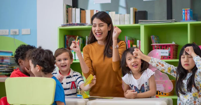 Image shows a teacher laughing with young children in a classroom
