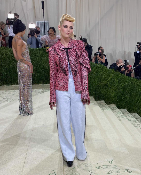 American actress Kristen Stewart stands on The Met red carpet. She is a light-skin-toned woman wearing a ruffled pink jacket and white pants. Her hair is pulled back in a retro ponytail style. Guests and photographers stand by a hedge in the background.