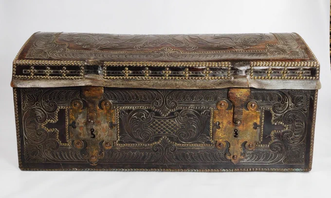 A dark rectangular trunk, with ornate designs in leather.  Two large metal latches are on the front. 