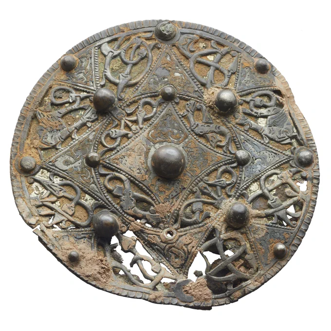 A circular, silver-and-copper coloured brooch decorated with swirling animal forms and interlocking geometric patterns. The rim is notched and there are small round balls affixed around the design.  