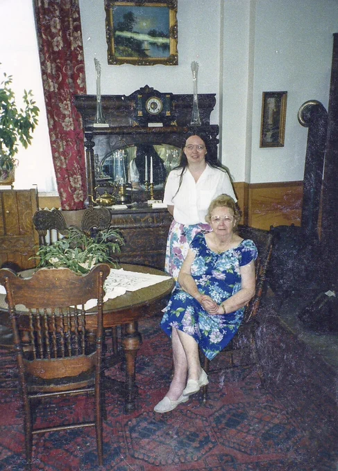 Cathy English stands behind Ruby Nobbs, museum manager, in a museum parlor exhibit featuring a round table, a buffet, damask curtains, and a patterned rug. The colour photograph was taken circa 1985. 