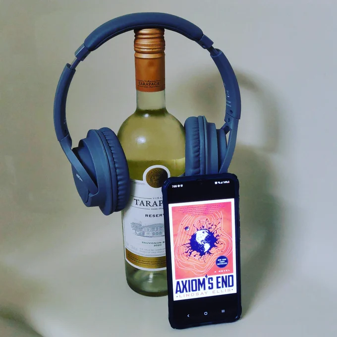 A bottle of wine wine with a pair of grey headphones balanced on top of it holding up a mobile phone with the cover of Axiom's End displayed. All are on a ledge in a hotel bathroom.