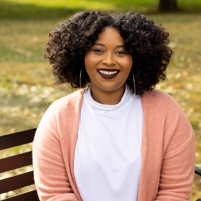 Photo of a smiling Dr. Devin Swiner sitting on a bench outside. She wears a light pink sweater, silver hoops, and a dark lipstick.