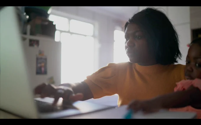 Woman types on a laptop with one hand while holding a child in the other arm.