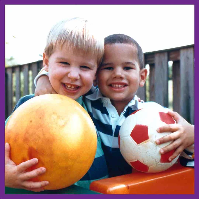 Two young boys with their arms round each other smiling at the camera. One holds a yellow ball while the other holds a traditional football with white and red hexagons. They stand in front of a fence.