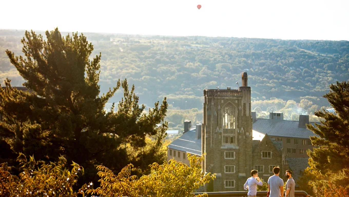 Three students stand at the Libe Slope overlook, with a grey-colored War Memorial and green and yellow trees in the background.
